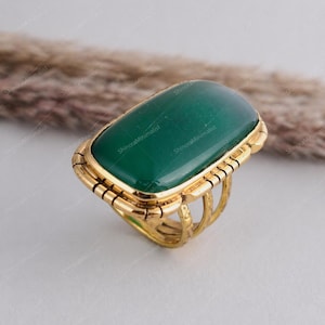 May include: A gold ring with a large, rectangular, emerald-green stone. The ring has a detailed, textured band and a decorative setting around the stone. The ring's design is elegant and eye-catching.