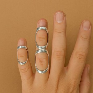 May include: Three silver rings are shown on fingers against a neutral background. The rings have an elongated oval shape, covering the top of the finger. The rings are made of a shiny, metallic material.