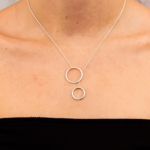 May include: A silver necklace with two linked open circles. The upper circle is bigger than the lower one. The necklace is suspended from a fine chain, creating a minimalist and modern jewellery piece.