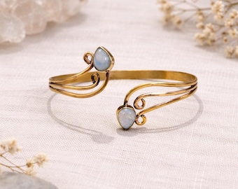 May include: Open cuff bracelet in a gold tone, featuring two pear-shaped, pale blue gemstones. The bracelet has an intricate design with curved lines and decorative swirls. The jewelry is displayed on a white, textured surface.