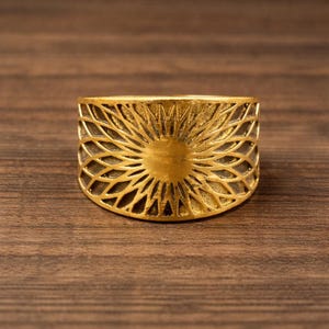 Op de afbeelding: Een goudkleurige ring met een zonnemotief. De ring heeft een centraal cirkelvormig element omgeven door een gedetailleerd, opengewerkt patroon. De ring staat op een houten achtergrond.