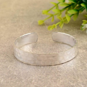 May include: A silver-colored cuff bracelet with a hammered texture. The open-ended bracelet is displayed on a neutral surface, with green foliage in the background. The bracelet has a simple, minimalist design.