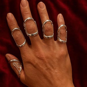 以下が含まれることがあります： 複数の銀色の指輪をつけた手。指輪は、開いた長方形や曲線など、ユニークなデザインです。ジュエリーは濃い赤色の背景に展示されており、金属的な輝きを際立たせています。指輪はモダンで芸術的です。