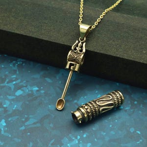 May include: A gold-colored pendant necklace with a detailed, ornate design. The pendant features a trident-like top and a cylindrical body with a small spoon-like attachment. A matching cylindrical container is also present.