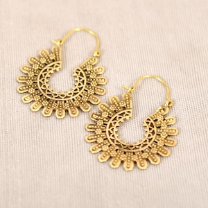 May include: A pair of gold-colored hoop earrings. Each earring features a sunburst design with intricate cut-out patterns. The earrings have a hook closure and are displayed on a neutral-colored surface. The earrings are a jewelry item.