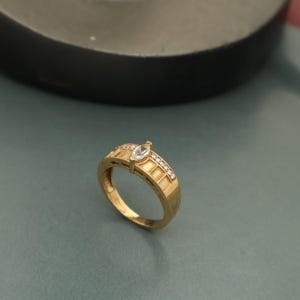 Puede incluir: Un anillo de oro con una piedra preciosa clara de talla marquesa en la parte superior y pequeñas piedras preciosas claras a lo largo de la banda. El anillo tiene un diseño texturizado en la banda. El anillo está sobre un fondo verde azulado apagado.