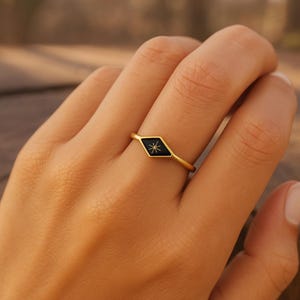 Puede incluir: Un anillo dorado con un centro negro en forma de diamante. El centro presenta un diseño de estrella blanca. El anillo es simple y elegante, adecuado para el uso diario. El anillo se muestra en un dedo.
