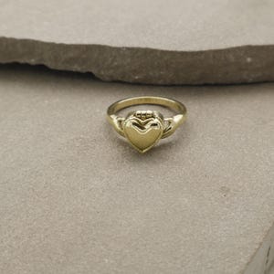 Puede incluir: Un anillo dorado con un diseño en forma de corazón. El anillo presenta una pieza central detallada en forma de corazón y una banda con elementos decorativos. El anillo está sobre un fondo neutro.