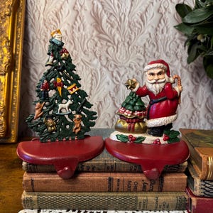 May include: Two Christmas stocking holders. One is a green Christmas tree with ornaments, the other is Santa Claus holding a gift and a Christmas tree. Both are red and sit on top of a stack of books.