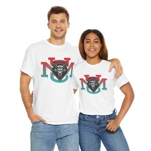 Puede incluir: Camisetas blancas con un gráfico de cabeza de lobo dentro de un diamante, con el texto "LOBOS" arriba y "NM" abajo. El diseño es en rojo, turquesa y gris. Las camisetas son usadas por dos personas.