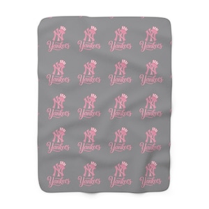 Op de afbeelding: Een rechthoekige grijze fleece deken met een herhalend patroon van het roze New York Yankees logo en het woord Yankees geschreven in een cursief lettertype. Het ontwerp is gelijkmatig verdeeld over het oppervlak van de zachte stof.