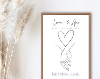 May include: A framed print featuring a minimalist line drawing of two hands holding each other, forming a heart shape. The names "Lorena & Alex" and the date "06. JUNI 2025" are above the hands. The phrase "HAND IN HAND EIN LEBEN LANG" is below.