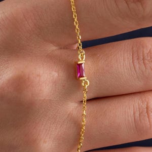 Könnte beinhalten: Ein zartes Goldkettenarmband mit einem einzelnen, kleinen, rechteckig geschliffenen fuchsiafarbenen Edelstein in einer Goldfassung. Die feine Gliederkette ist poliert und glänzend, was den Kontrast zwischen dem Goldmetall und dem Stein hervorhebt.