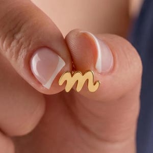 May include: A small gold-toned stud earring shaped like the lowercase letter m in a cursive script font. The earring has a smooth, polished finish and is held between two fingers, showcasing the letter's distinct curves and gold color.