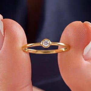 May include: A delicate gold ring with a small, round, clear gemstone set in a bezel. The ring has a thin band and a polished finish. The ring is held between two fingers against a dark blue background.