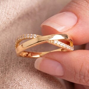 May include: A gold ring with a crisscross design. One band is encrusted with small, clear gemstones. The ring is held against a neutral-toned fabric background. The ring is a piece of jewellery.