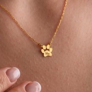 May include: A gold necklace featuring a fine chain and a paw print pendant. The pendant has a small inscription. The necklace is worn around the neck.