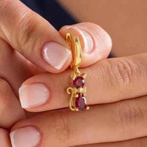 May include: A gold-colored cat-shaped earring with a red gemstone body and a bow. The earring is held up by a gold hoop. The cat's head and body are made of red gemstones.