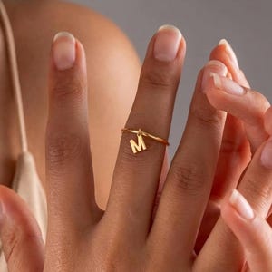 Könnte beinhalten: Ein goldfarbener Ring mit einem zarten Band und einem hängenden Buchstaben "M". Der Ring wird an einem Finger getragen, wodurch sein schlichtes, aber elegantes Design hervorgehoben wird. Der Ring ist ein personalisiertes Schmuckstück.