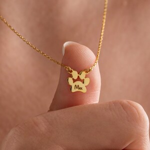 May include: A gold-coloured necklace with a paw print pendant. The pendant has a small bow detail and the name "Max" engraved within the paw print. The necklace is displayed on a person's finger.