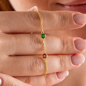 May include: A delicate gold chain bracelet featuring two colorful gemstones. The bracelet has a green and a red gemstone. The bracelet is held up by a person's hand.