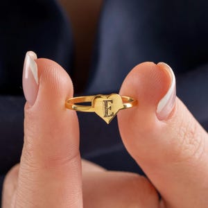 Könnte beinhalten: Ein goldfarbener Ring mit einem herzförmigen Mittelstück, in dessen Mitte der Großbuchstabe E eingraviert ist. Das Band ist dünn und metallisch und weist eine polierte Oberfläche auf, die das Licht reflektiert. Das Design ist schlicht.