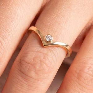 May include: A gold-colored ring with a V-shaped design and a small, round, clear gemstone at the point of the V. The ring is on a finger, and the background is a blurred view of skin and fabric.