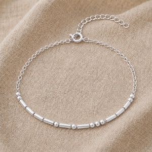 Könnte beinhalten: Ein silberfarbenes Kettenarmband mit einer Abfolge von abwechselnden kleinen runden Perlen und kurzen zylindrischen Metallröhrchen. Das Armband besteht aus einer feinen Gliederkette, einem runden Federringverschluss und einer verstellbaren Verlängerungskette.