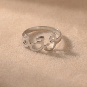 Op de afbeelding: Een zilveren ring met het nummer 333 uitgesneden in het midden. De ring is gemaakt van een glanzend metaal en ligt op een neutraal gekleurd oppervlak. De ring is een eenvoudige band met de nummers als belangrijkste ontwerpelement.