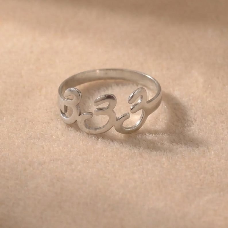 Op de afbeelding: Een zilveren ring met het nummer 333 uitgesneden in het midden. De ring is gemaakt van een glanzend metaal en ligt op een neutraal gekleurd oppervlak. De ring is een eenvoudige band met de nummers als belangrijkste ontwerpelement.