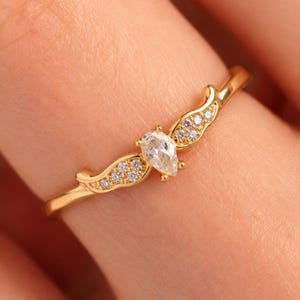 May include: A gold ring featuring a central pear-shaped clear stone flanked by two wing-shaped accents, each set with four small round clear stones. The band is thin and polished gold, creating a delicate appearance with symmetrical detailing.