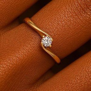 May include: A gold ring featuring a single round brilliant-cut clear gemstone set in a bypass-style band. The metal band curves around the central stone, creating a smooth, continuous silhouette that highlights the clarity of the gemstone.