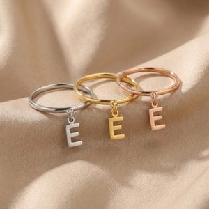 Könnte beinhalten: Drei dünne Metallringe, jeder mit einem kleinen baumelnden Buchstaben-Anhänger E. Das Set enthält einen silberfarbenen Ring, einen goldfarbenen Ring und einen roségoldfarbenen Ring, alle mit schlichtem Banddesign und hängendem Initial-Anhänger.