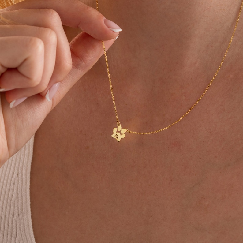 May include: A delicate gold necklace with a paw print pendant. The pendant is a small, detailed representation of a paw. The necklace is shown against a skin tone background, highlighting the jewelry's design and colour.