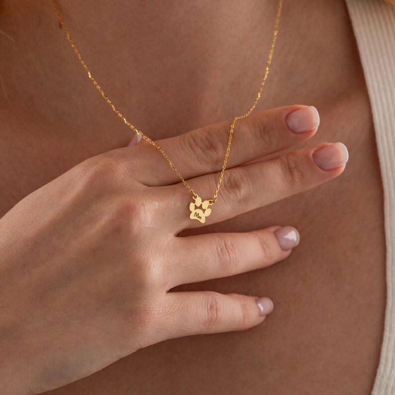 May include: A gold-coloured necklace featuring a paw print pendant. The pendant has a small inscription. The necklace is displayed against a neutral background.