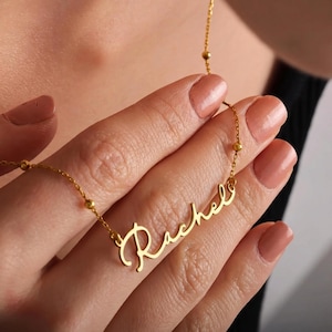 May include: A gold-colored necklace with the name "Rachel" in cursive script. The necklace is held by a person with manicured nails. The chain has small, round beads. The necklace is a personalized jewelry item.