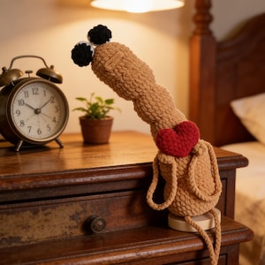 May include: A handmade, tan-colored crochet doll with a red heart and black pom-pom eyes. The doll is sitting on a wooden nightstand next to a vintage alarm clock and a small potted plant. The doll's legs dangle down.