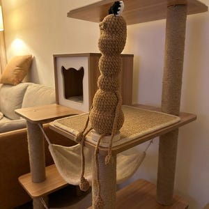 May include: A multi-level cat tree with wooden platforms, sisal-wrapped posts, and a beige hammock. A handmade, brown crocheted figure with dangling limbs sits on the top platform. The cat tree is in a room with a neutral color scheme.