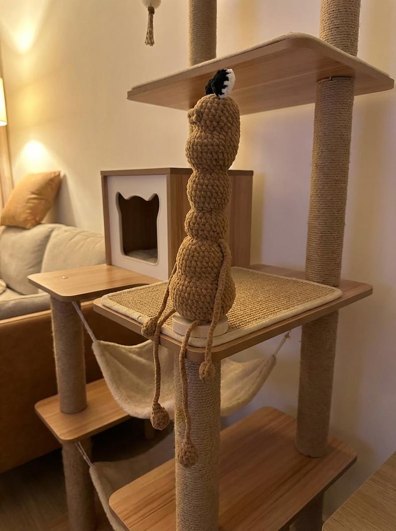 May include: A multi-level cat tree with wooden platforms, sisal-wrapped posts, and a beige hammock. A handmade, brown crocheted figure with dangling limbs sits on the top platform. The cat tree is in a room with a neutral color scheme.