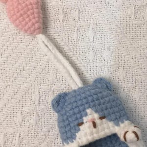 May include: A crocheted blue and white cat-shaped pouch featuring a small pink crocheted fish attached by a white yarn cord. The cat design includes two small ears, a white face patch, and two white paws with brown embroidered details on the front.