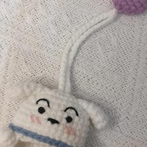 May include: A white crocheted animal head with black embroidered eyes, arched eyebrows, and pink stitched cheeks. It features a blue collar, a long white crocheted cord extending upward, and a small purple crocheted heart attached to the end.