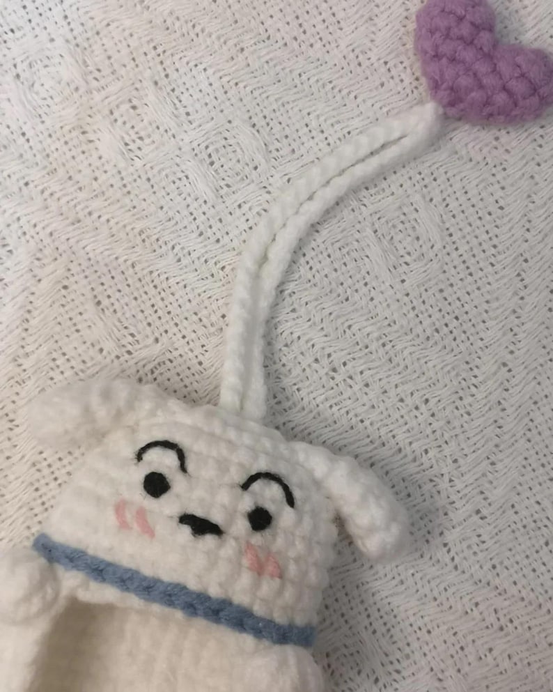 May include: A white crocheted animal head with black embroidered eyes, arched eyebrows, and pink stitched cheeks. It features a blue collar, a long white crocheted cord extending upward, and a small purple crocheted heart attached to the end.