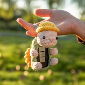 May include: A crocheted key fob cover shaped like a turtle, featuring a yellow hat, a light green shell, and a beige body. The key fob is visible within the turtle's body. The turtle has black eyes and a smiling mouth. The background is blurred green grass.