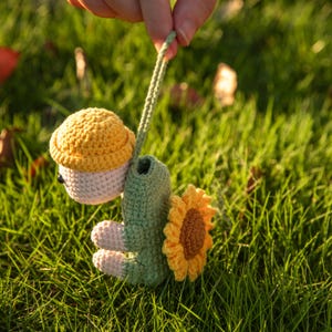 May include: A crocheted turtle figurine featuring a light pink head, a yellow textured shell, and a green body. The turtle has a large yellow sunflower attached to its back and a long green crocheted loop extending from the top of its head.