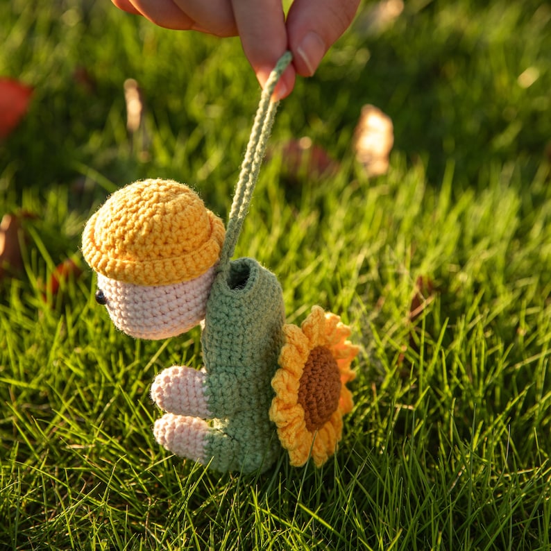 May include: A crocheted turtle figurine featuring a light pink head, a yellow textured shell, and a green body. The turtle has a large yellow sunflower attached to its back and a long green crocheted loop extending from the top of its head.