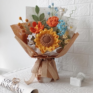 May include: A bouquet of crocheted flowers featuring a large yellow sunflower, orange blossoms, blue sprigs, and green leaves. The arrangement is wrapped in brown textured paper with a matching ribbon tie, creating a decorative floral display.