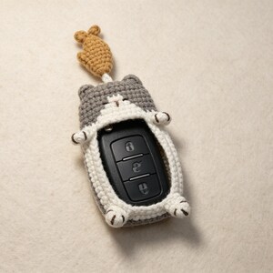 May include: A gray and white crocheted cat-shaped key fob cover featuring a small tan fish charm attached to the top. The cover encases a black rectangular car key remote with three buttons, showing the cat's ears, paws, and tail details.