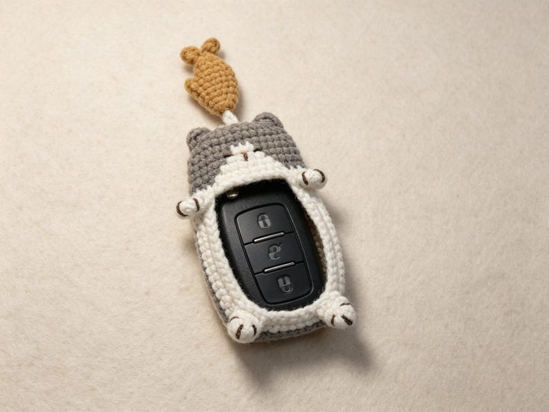 May include: A gray and white crocheted cat-shaped key fob cover featuring a small tan fish charm attached to the top. The cover encases a black rectangular car key remote with three buttons, showing the cat's ears, paws, and tail details.