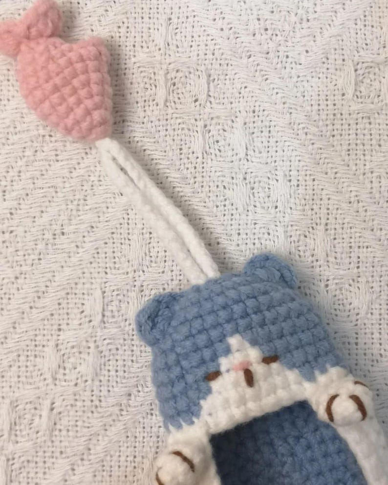 May include: A crocheted blue and white cat-shaped pouch featuring a small pink crocheted fish attached by a white yarn cord. The cat design includes two small ears, a white face patch, and two white paws with brown embroidered details on the front.