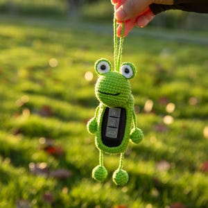 May include: A bright green crocheted frog key fob cover featuring a round head with white and black eyes, thin arms and legs with small round hands and feet, and a central opening that reveals a black car key fob with three silver buttons.